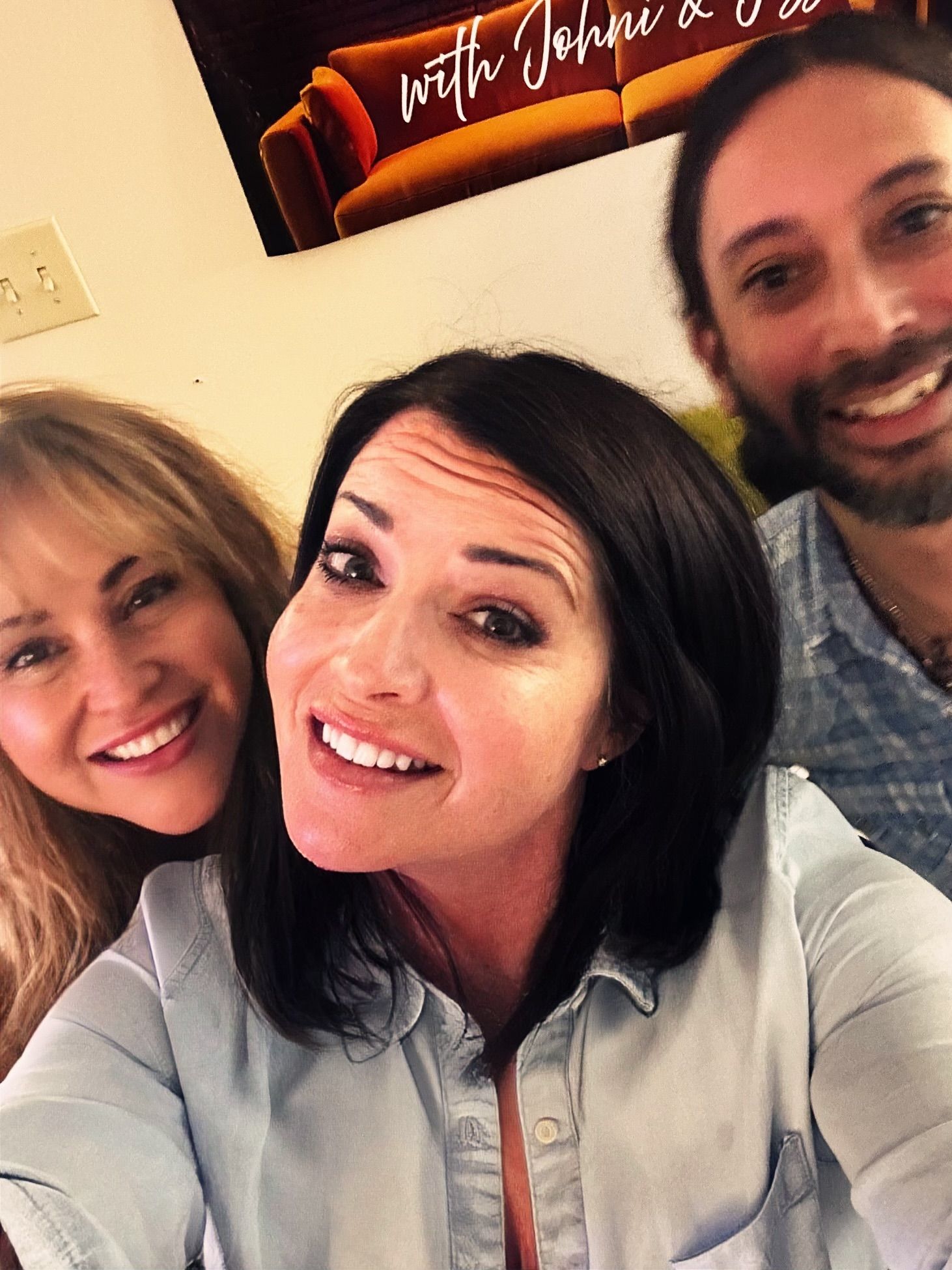 Three smiling people posing indoors, with a framed sign in the background.