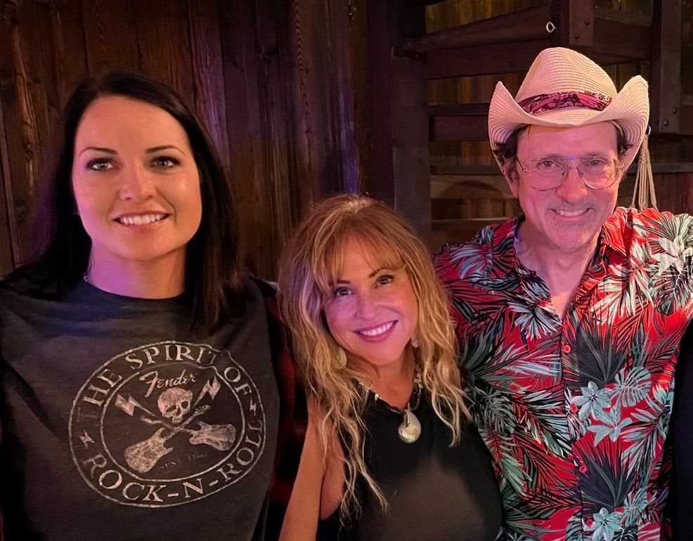Three people smiling in a dimly lit bar; one wears a cowboy hat and red floral shirt.