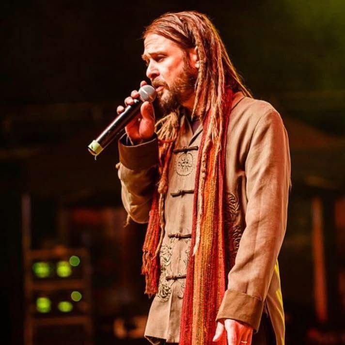 Singer with long dreadlocks performing into a microphone on a dimly lit stage