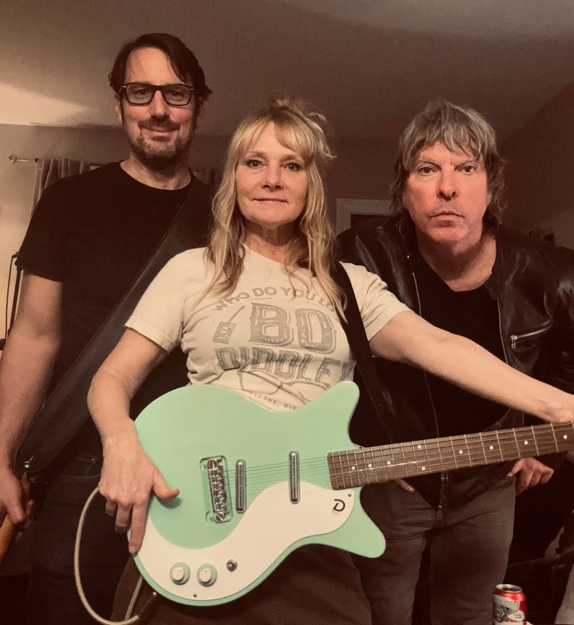 Three people posing with a mint-green electric guitar indoors.