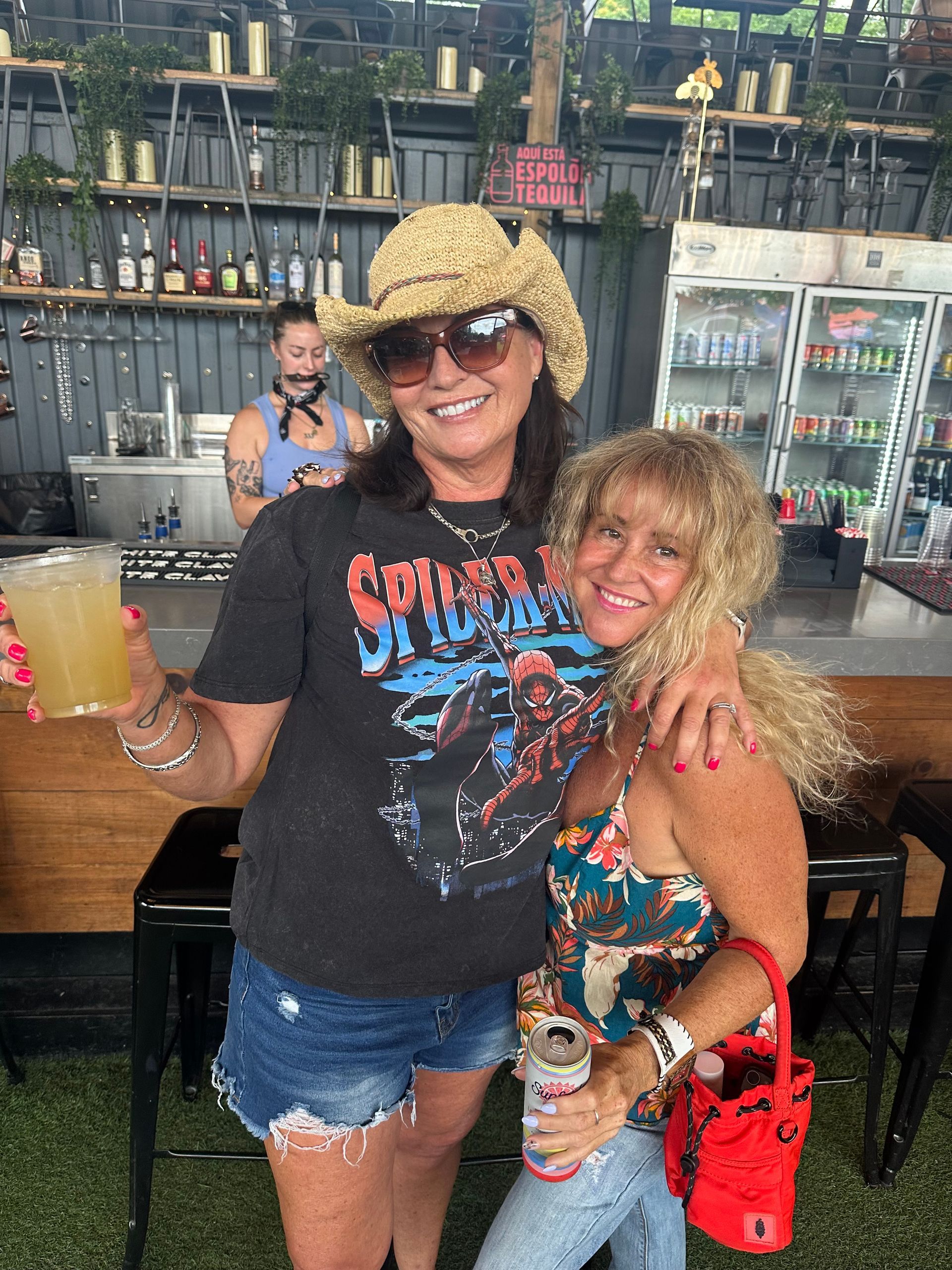 Two friends smiling at a bar, one in a hat and black graphic tee, the other in a red top and denim shorts.