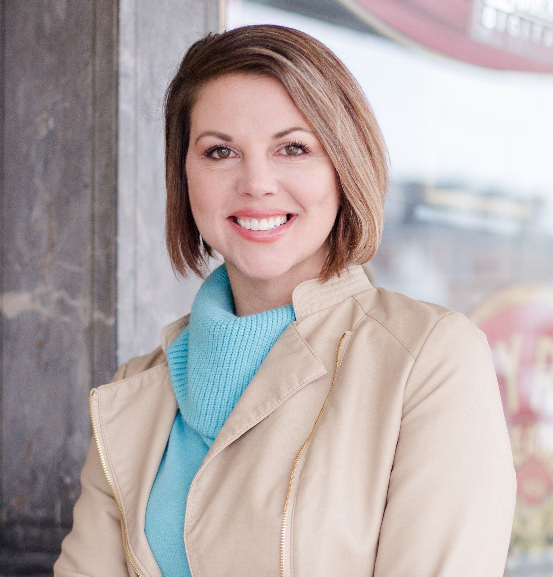 Smiling woman in beige jacket and blue sweater standing outdoors by a concrete wall