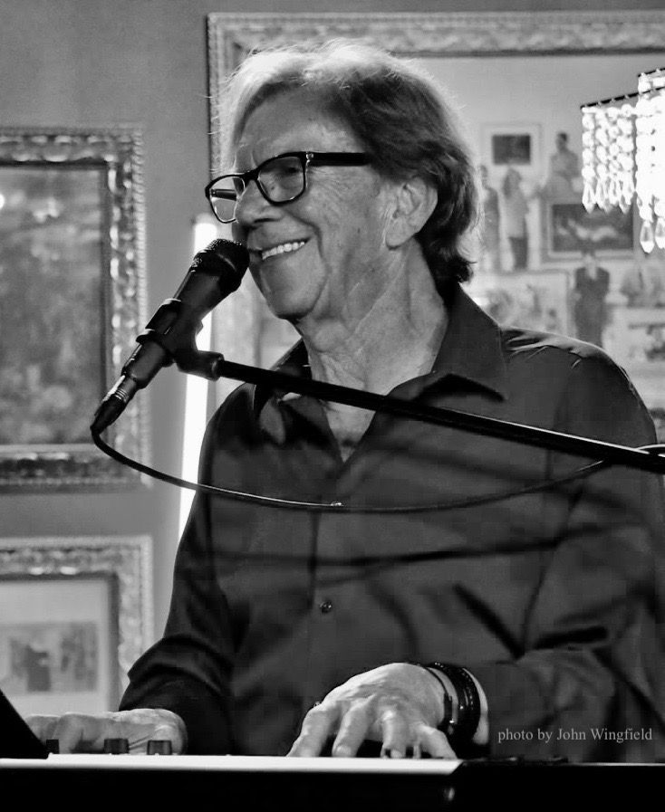 Musician smiling while singing into a microphone and playing keyboard in a framed, indoor setting