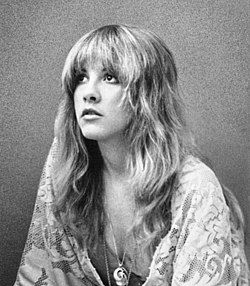 Stevie Nicks, in a lace shawl, looks upward with long wavy hair and a pendant.