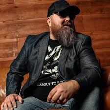 Bearded man in a black cap and jacket sitting against a wooden wall, looking to the side