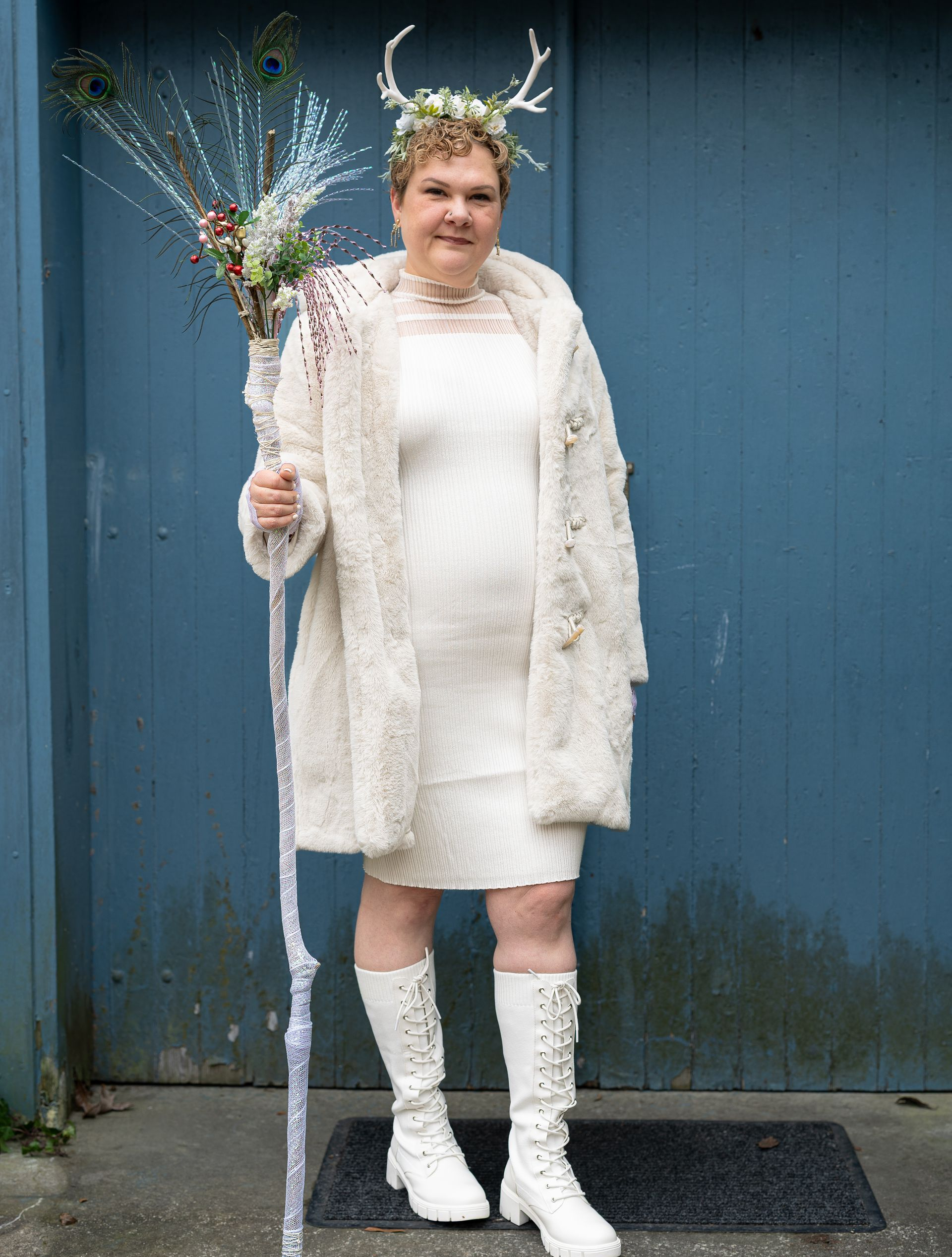 Person in a white outfit and antler headpiece holds a bouquet in front of a blue wall.