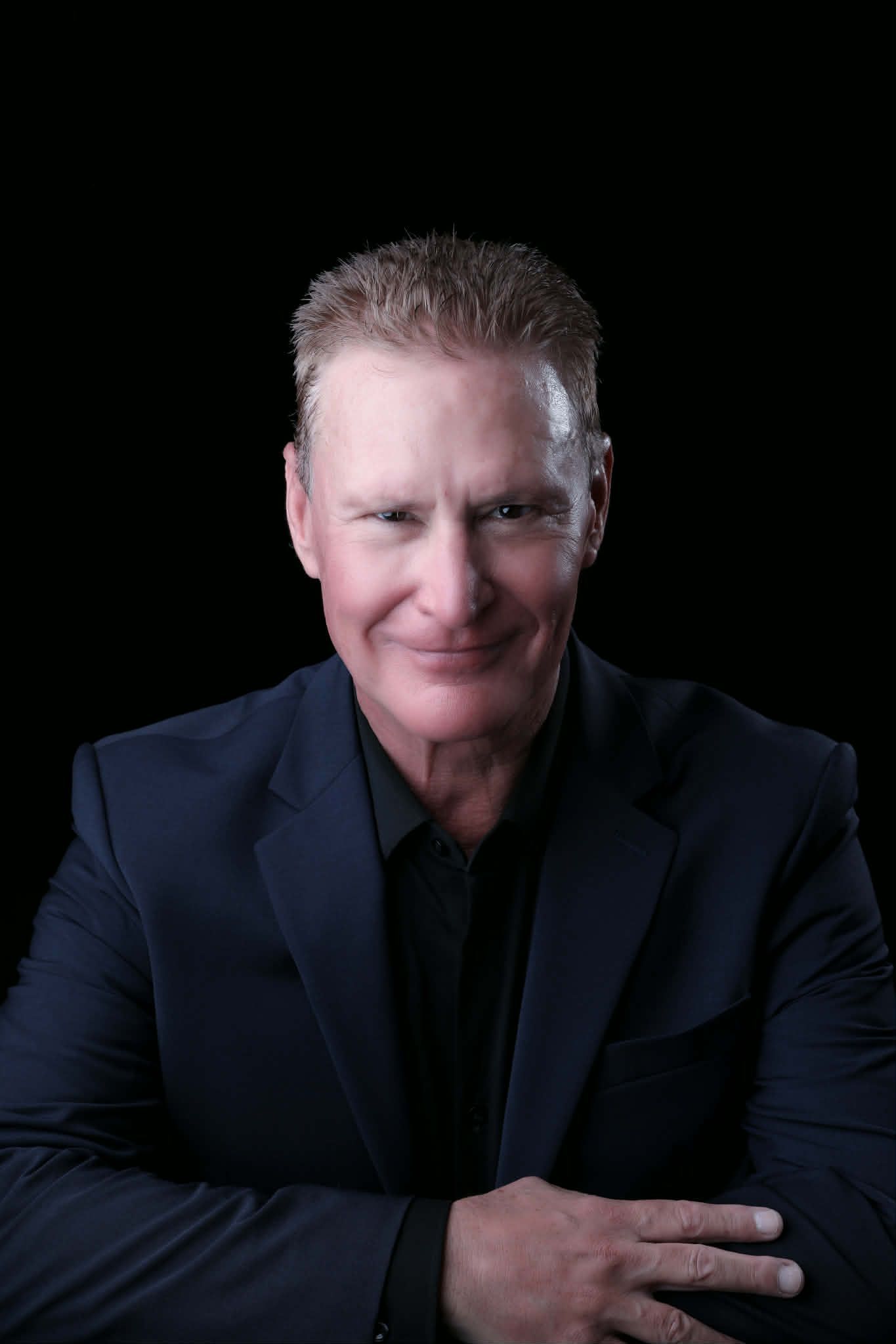 A person with light hair, wearing a dark suit and shirt, smiles while leaning forward against a plain black background.