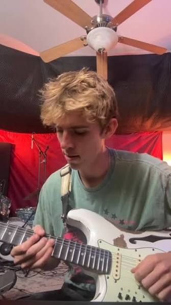 Person playing electric guitar in a room with red and black hanging fabric and a ceiling fan