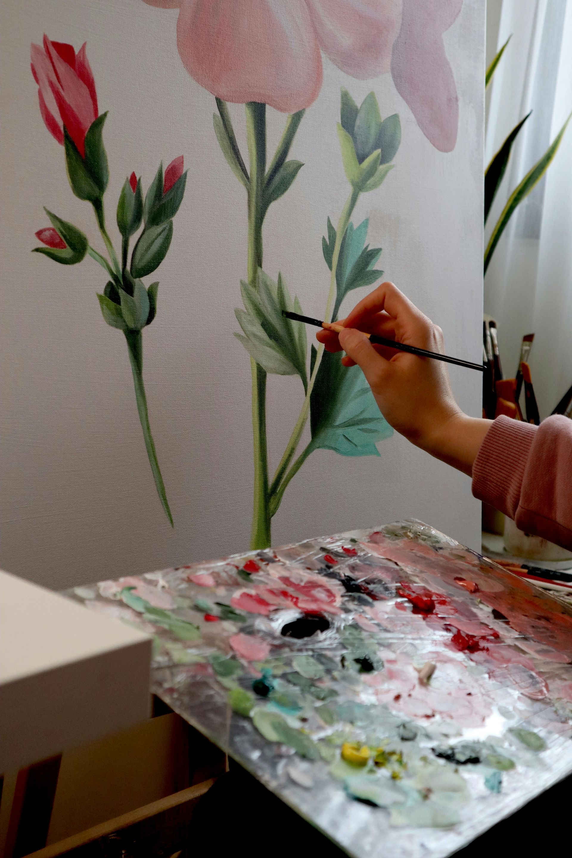 A person painting leaves on a floral canvas with a palette of mixed paint colors.