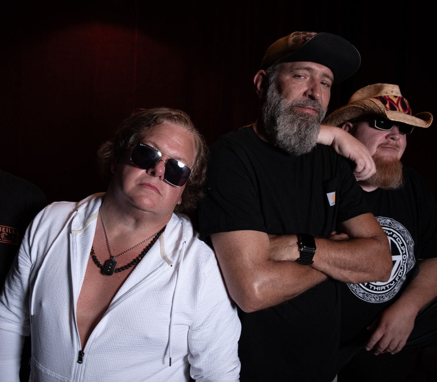 Four people posing against a black background, two with hats and one in sunglasses and a white shirt.