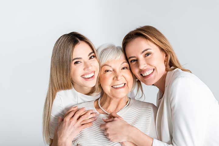 three generation of positive women smiling while looking at camera and hugging isolated on grey 
