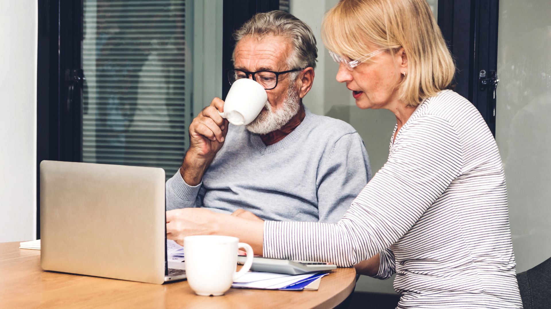 An elderly couple is sitting at a table drinking coffee and looking at a laptop.