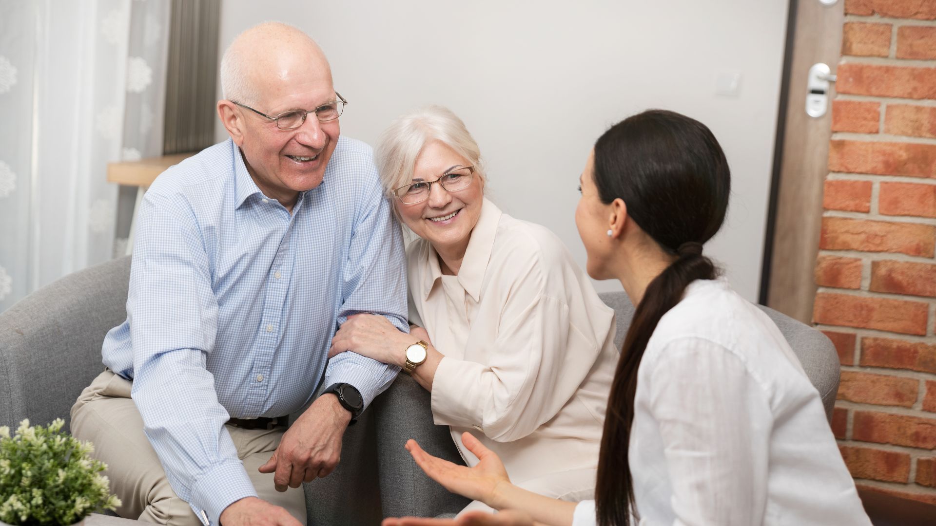 An elderly couple is sitting on a couch talking to a nurse.