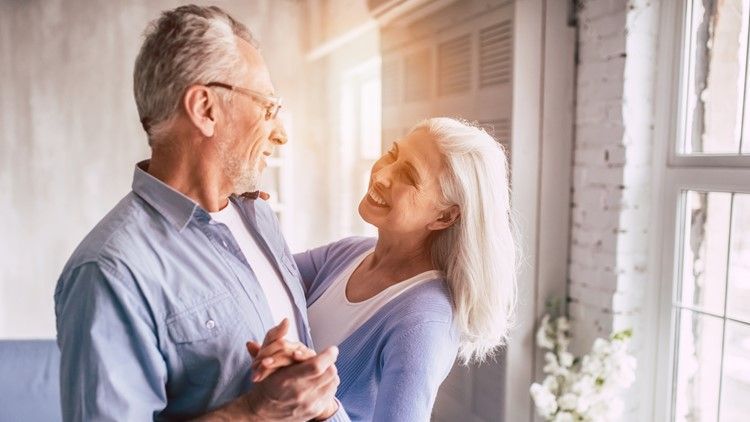 An elderly couple is dancing together in front of a window.