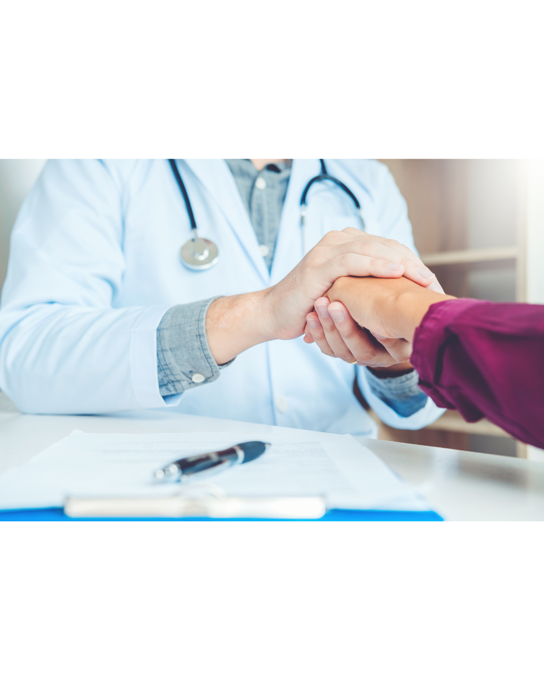 Doctor holding a patient's hand for comfort, stethoscope on shoulders, sitting at a desk.