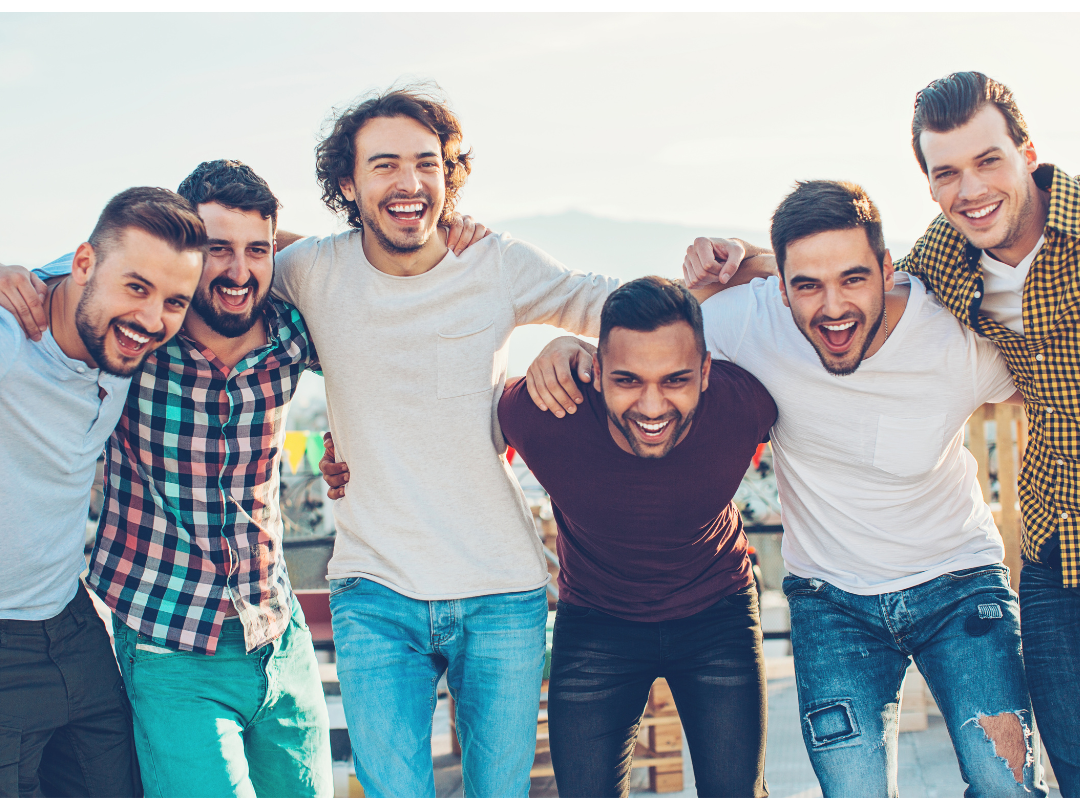 Group of seven men with arms around each other, smiling and laughing outdoors.