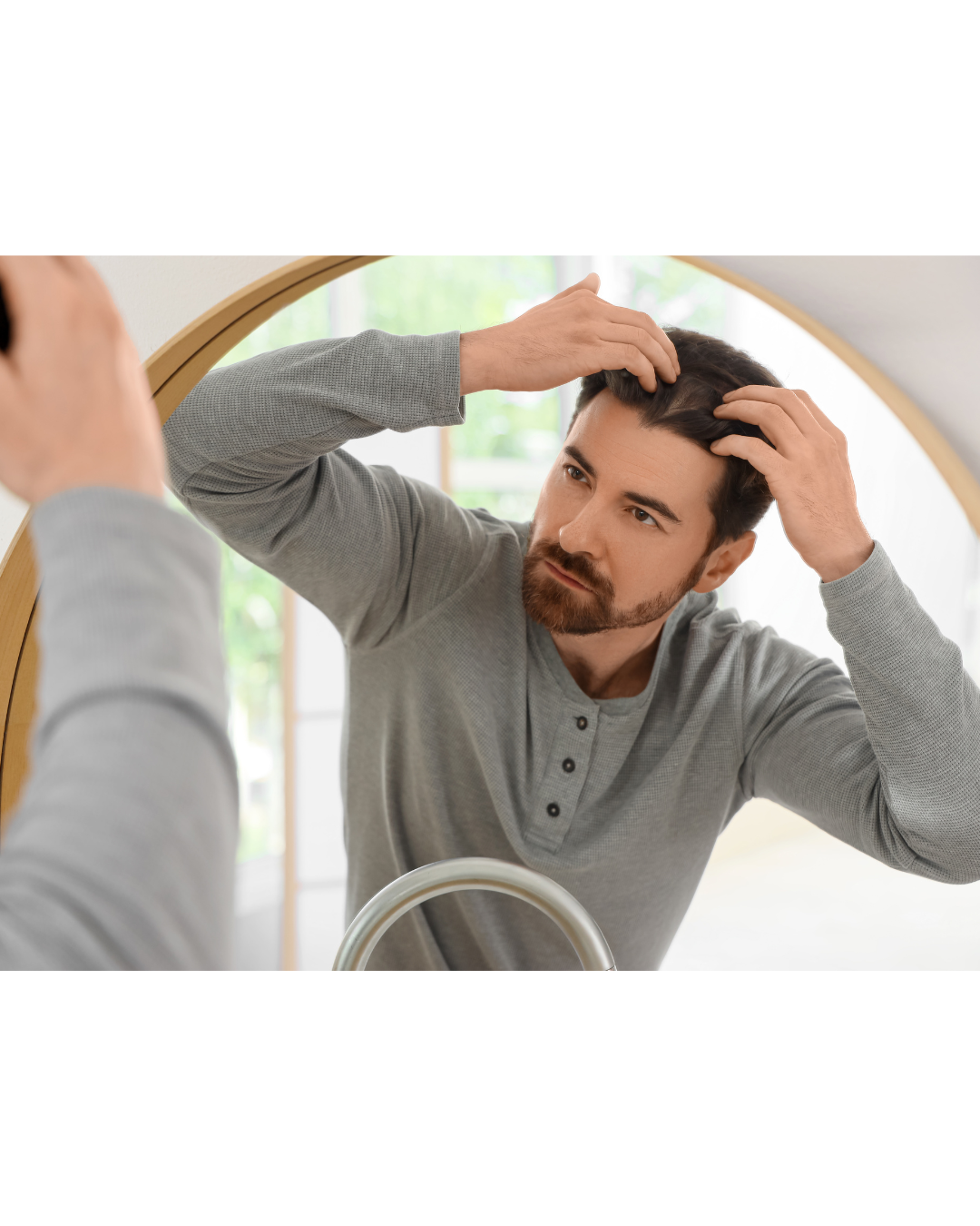 Man in gray shirt examining his hair in a bathroom mirror.