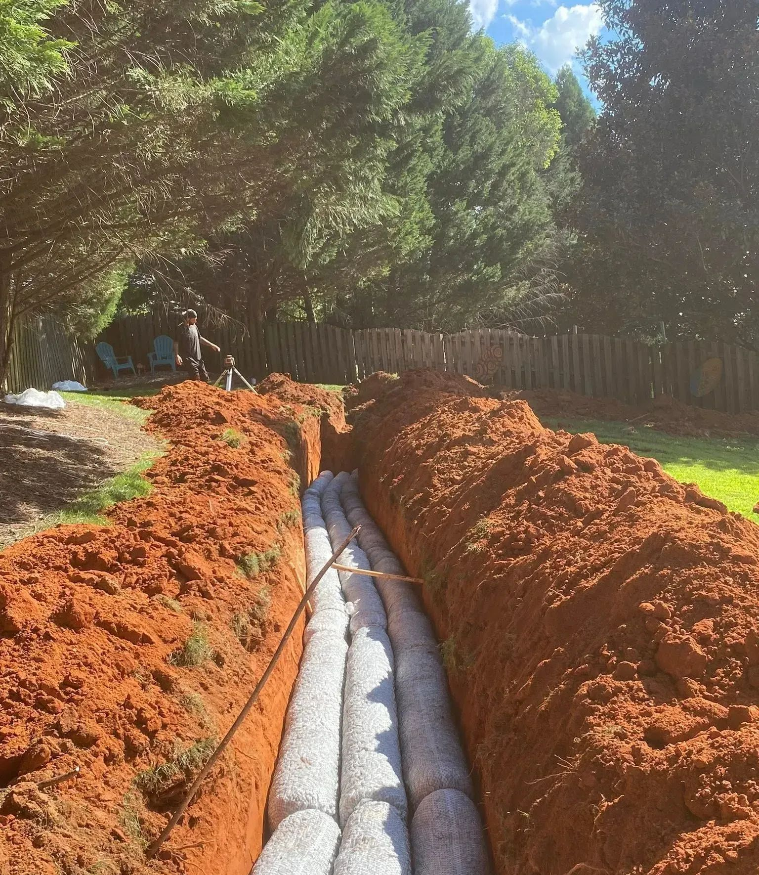 Trenched yard with orange soil walls and a gray drainage pipe being installed outdoors.