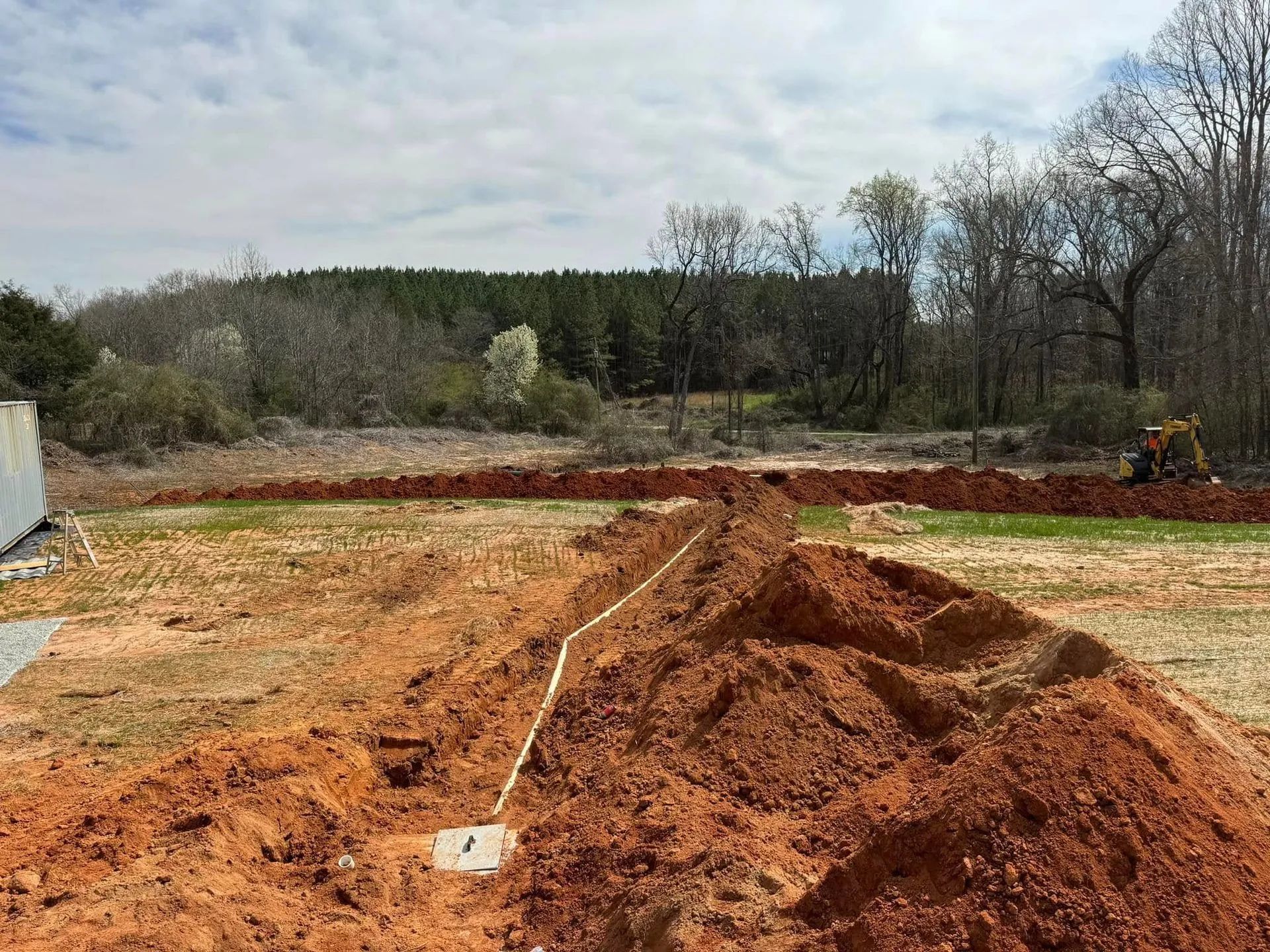Dirt road and red clay excavation site with piles of earth, a wooded treeline, and a cloudy sky