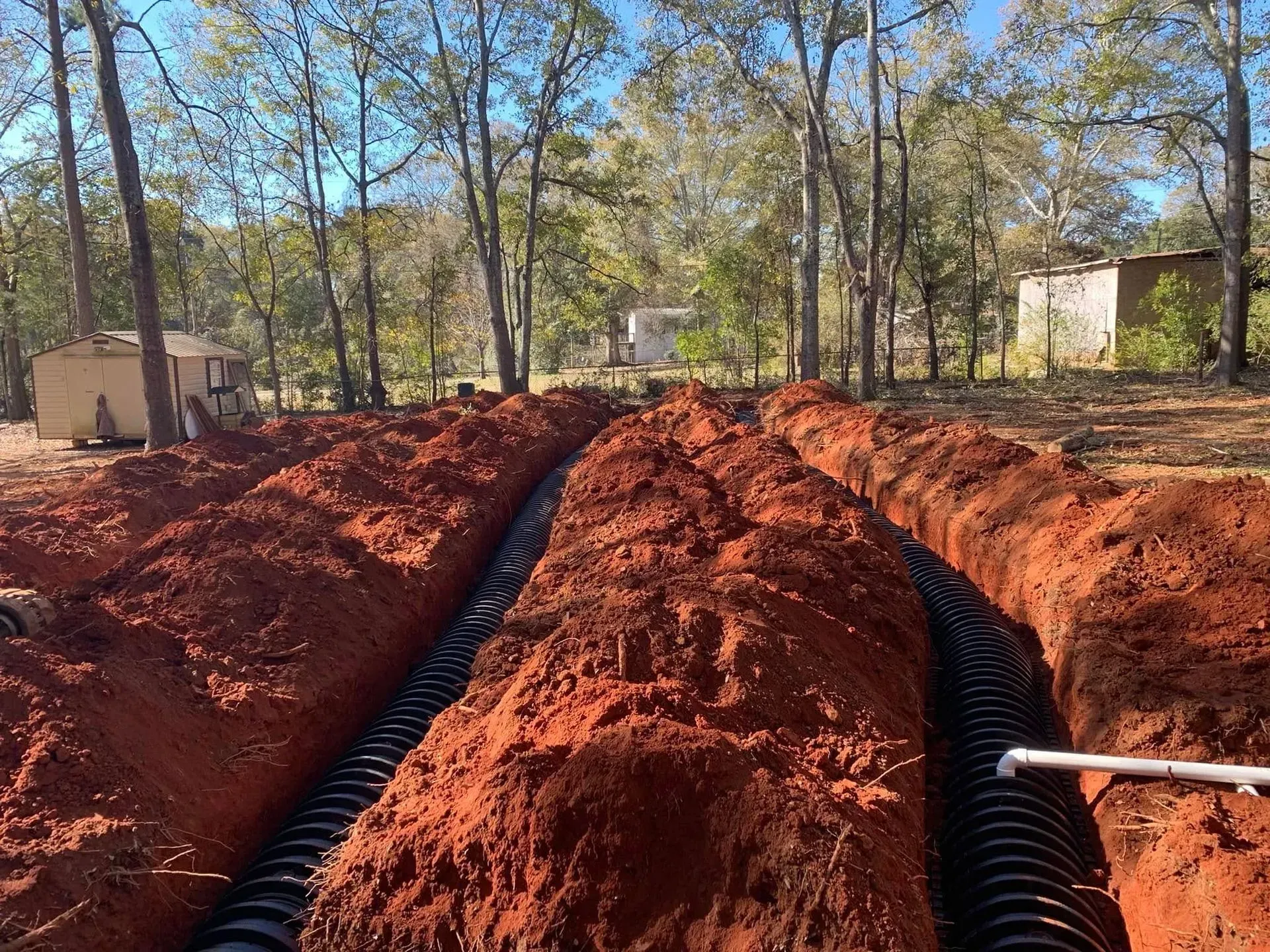 Trenches dug in red earth with drainage pipes installed, outdoors in a wooded area.
