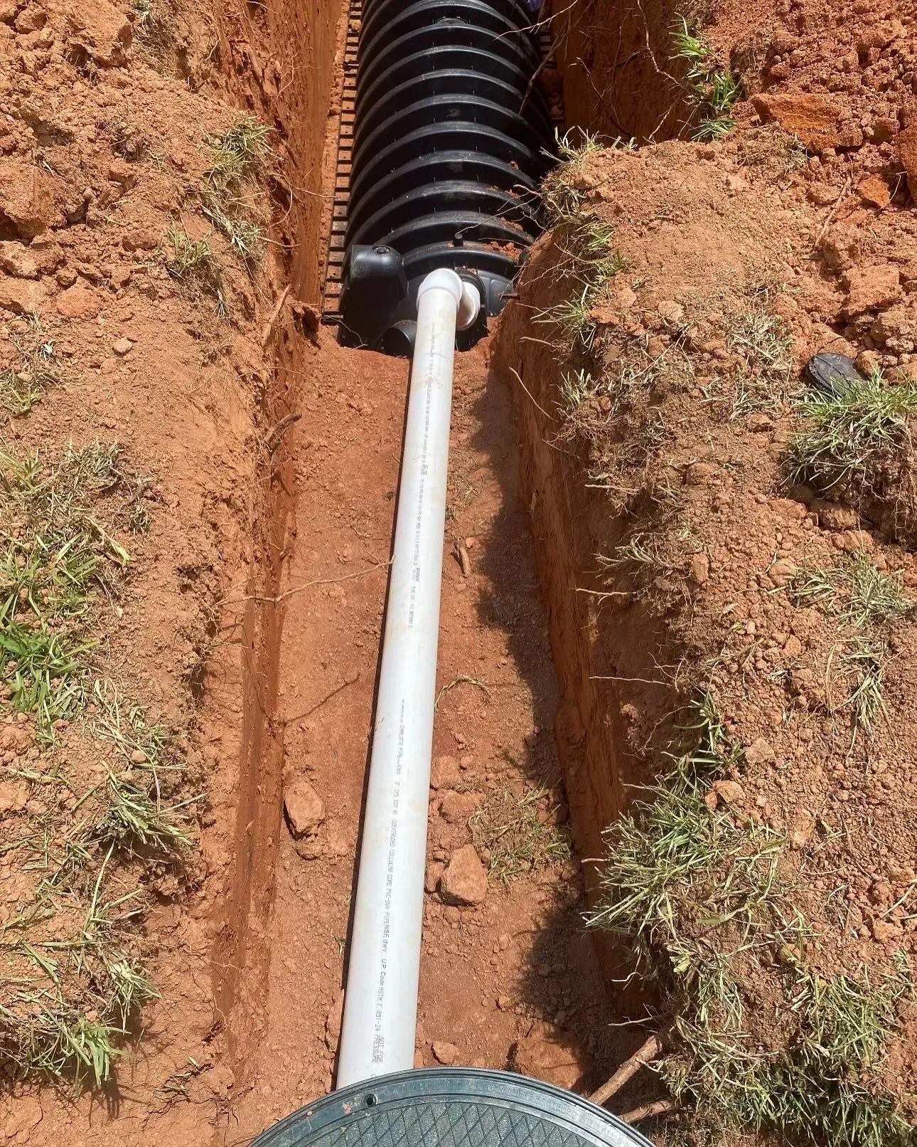 Drainage pipe and corrugated drain in a narrow trench in red soil. Grass at edge.
