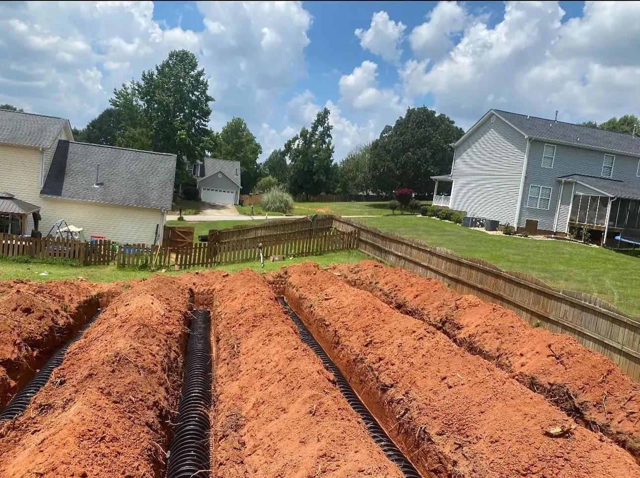 Rows of dug trenches in red soil, with houses and a fence in the background under a blue, cloudy sky.