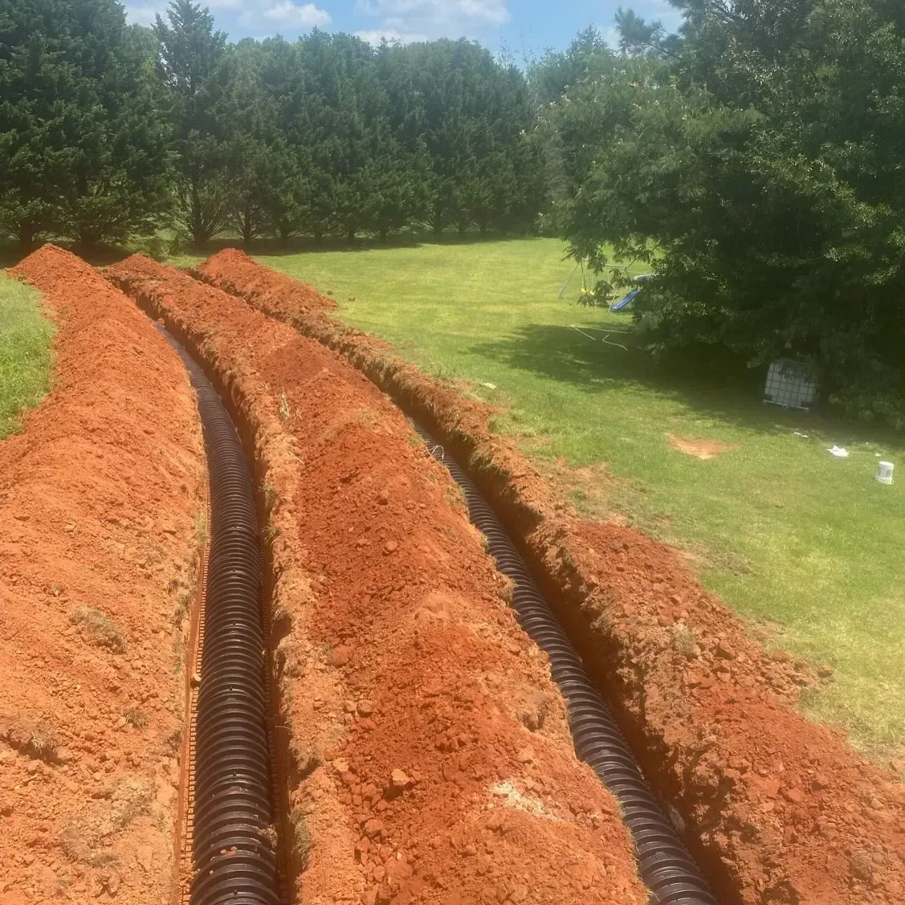 Trenches dug in a grassy yard, containing black corrugated drainage pipes, under a blue sky.