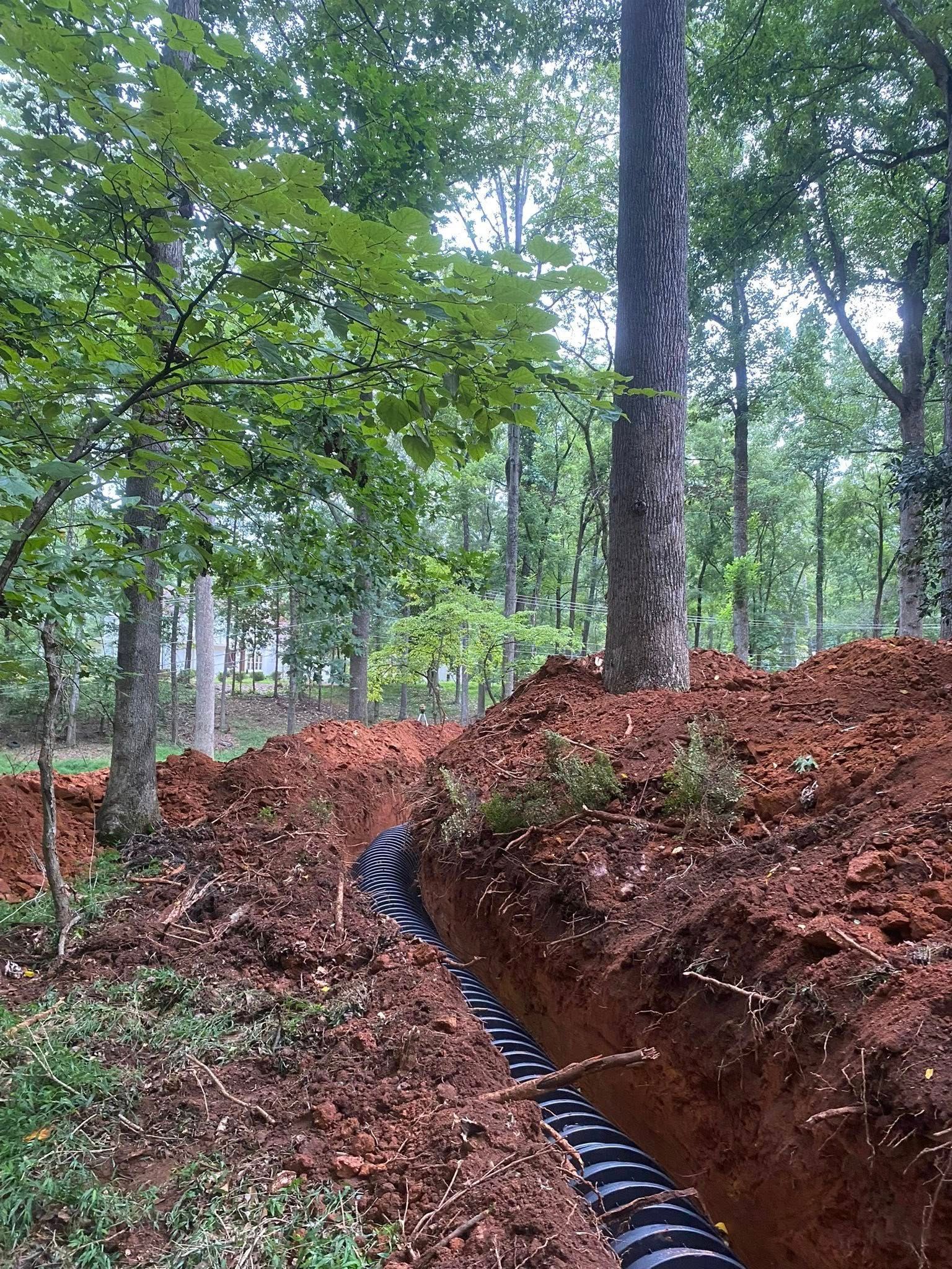 Trench in forest with black drainage pipe; red soil. Trees in background, cloudy sky.