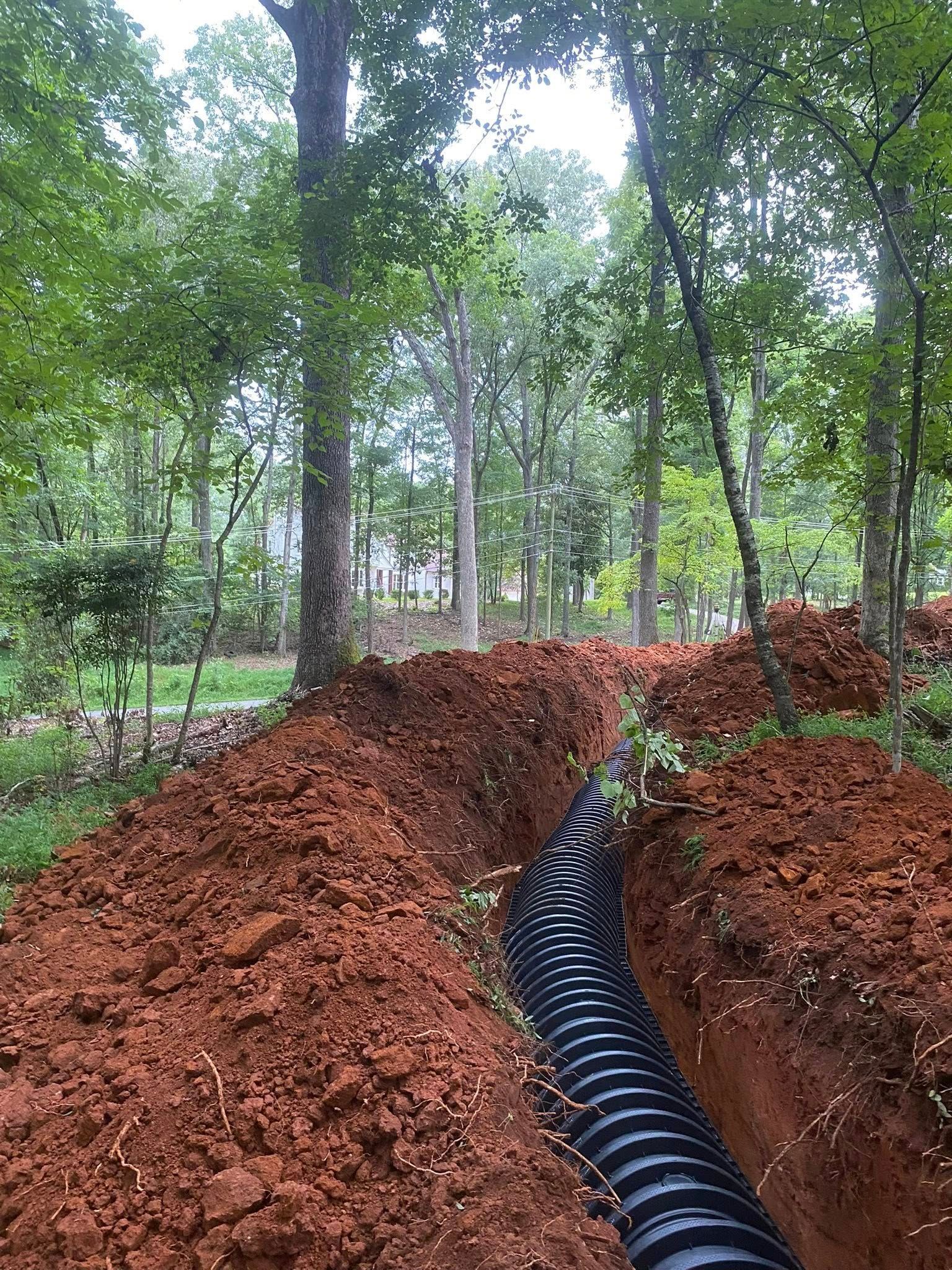 Drainage pipe in a trench of red soil, surrounded by trees in a wooded area.