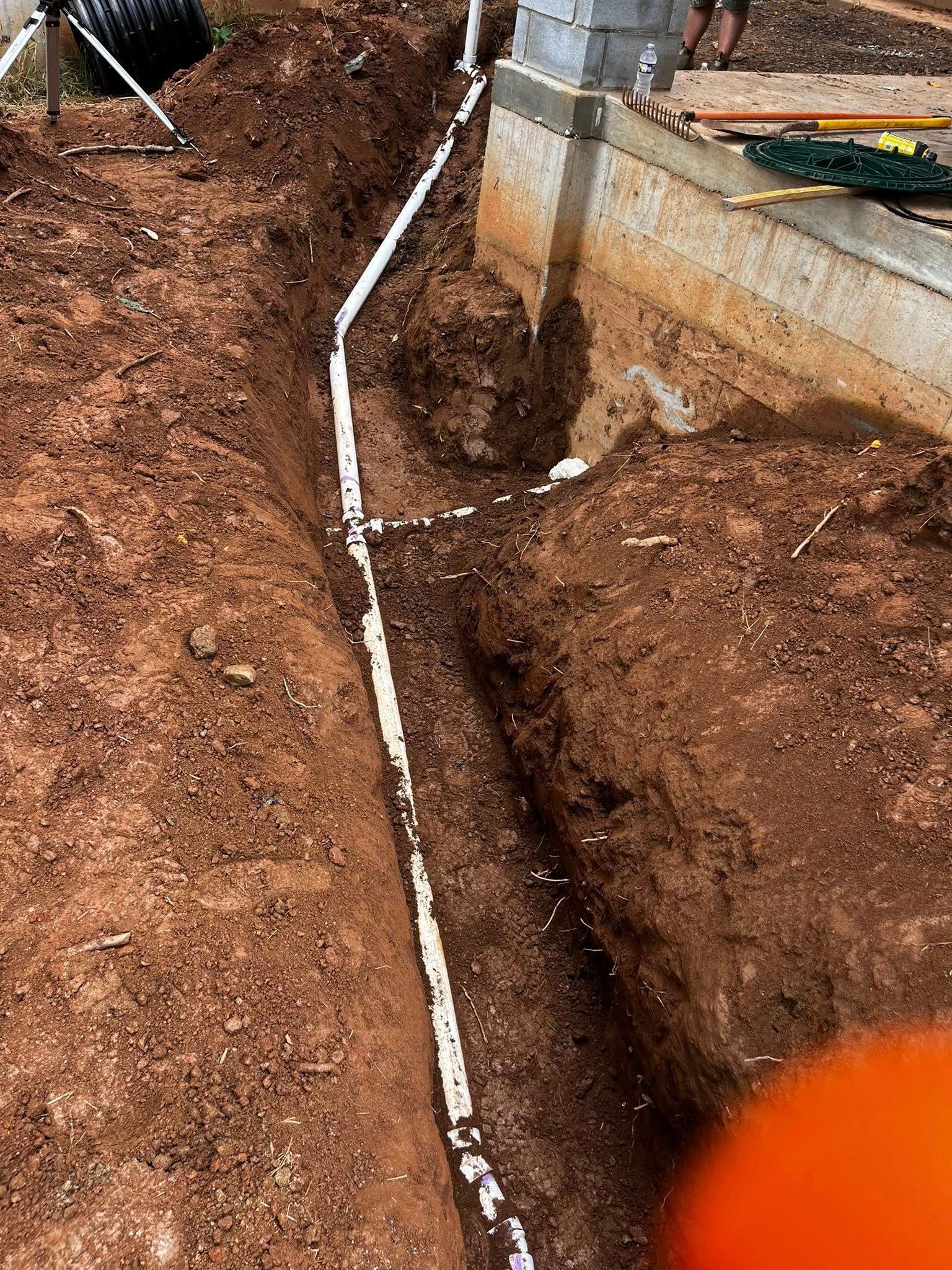 Trench dug in brown soil with white PVC pipes laid inside, near a concrete structure.