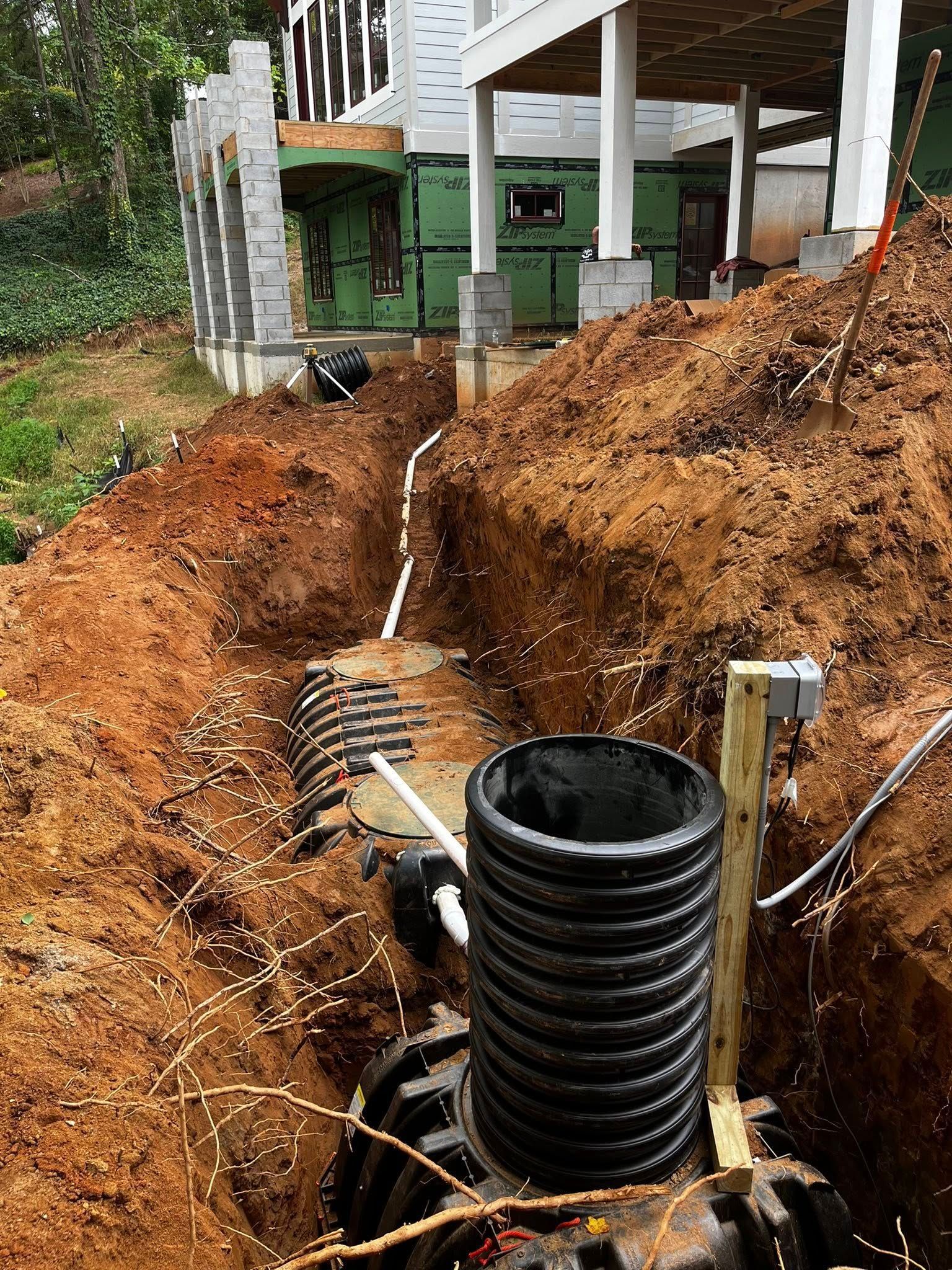 Construction site with an open trench containing pipes, a septic tank, and the foundation of a building.