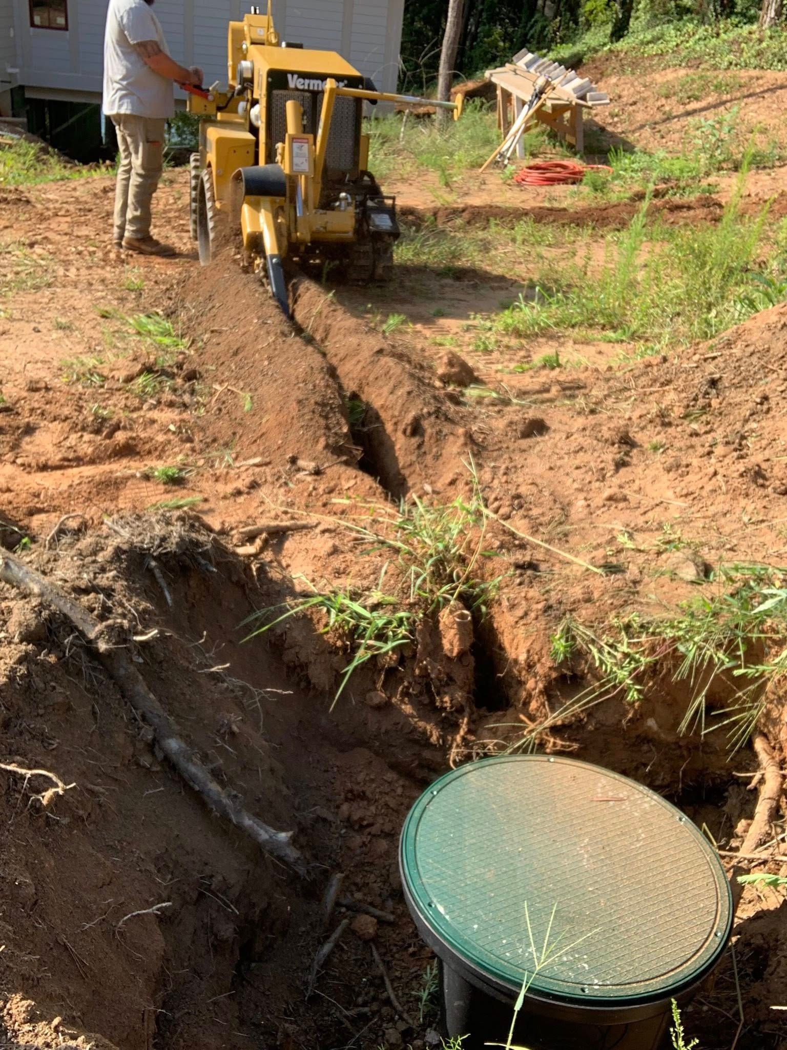 Man operating a yellow trencher digging a trench near a septic tank lid outdoors.