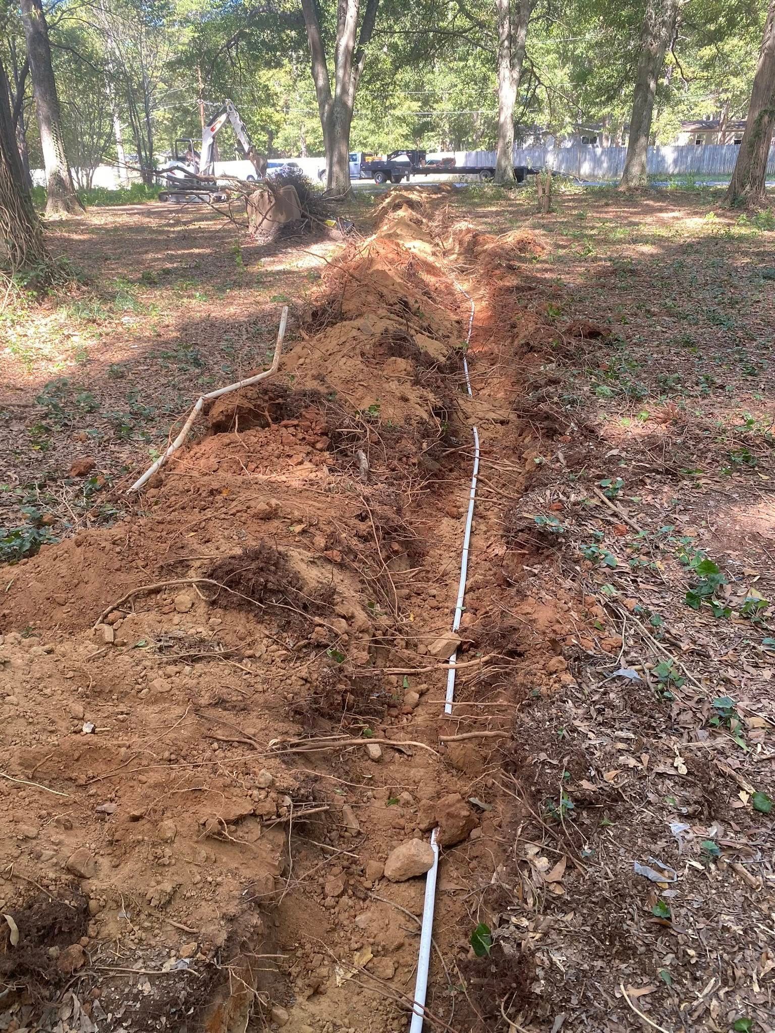 Trench dug in soil with white pipes running through it, in a wooded outdoor area.