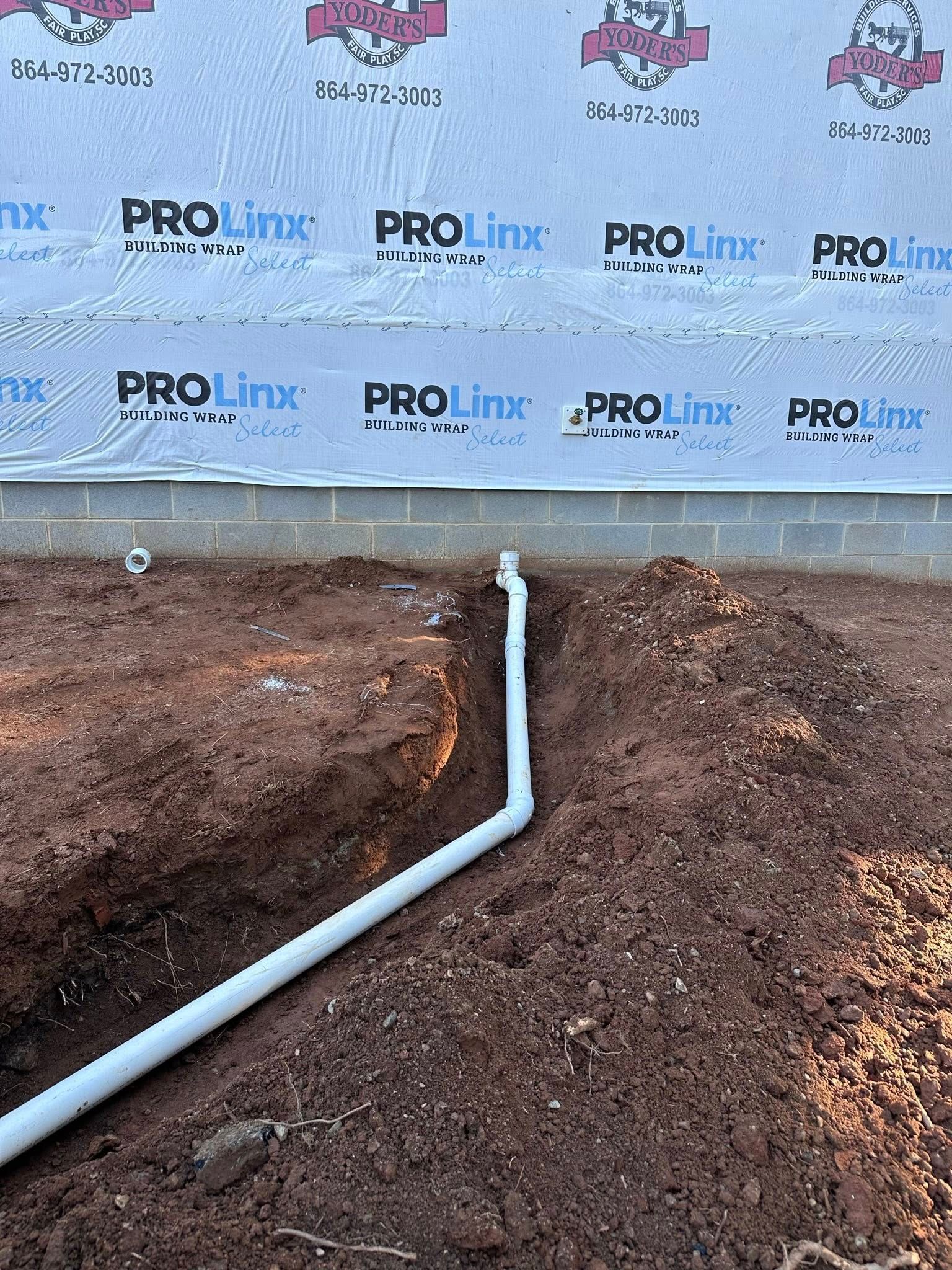 White PVC pipe in a dirt trench next to a foundation wall with blue and white wrap.