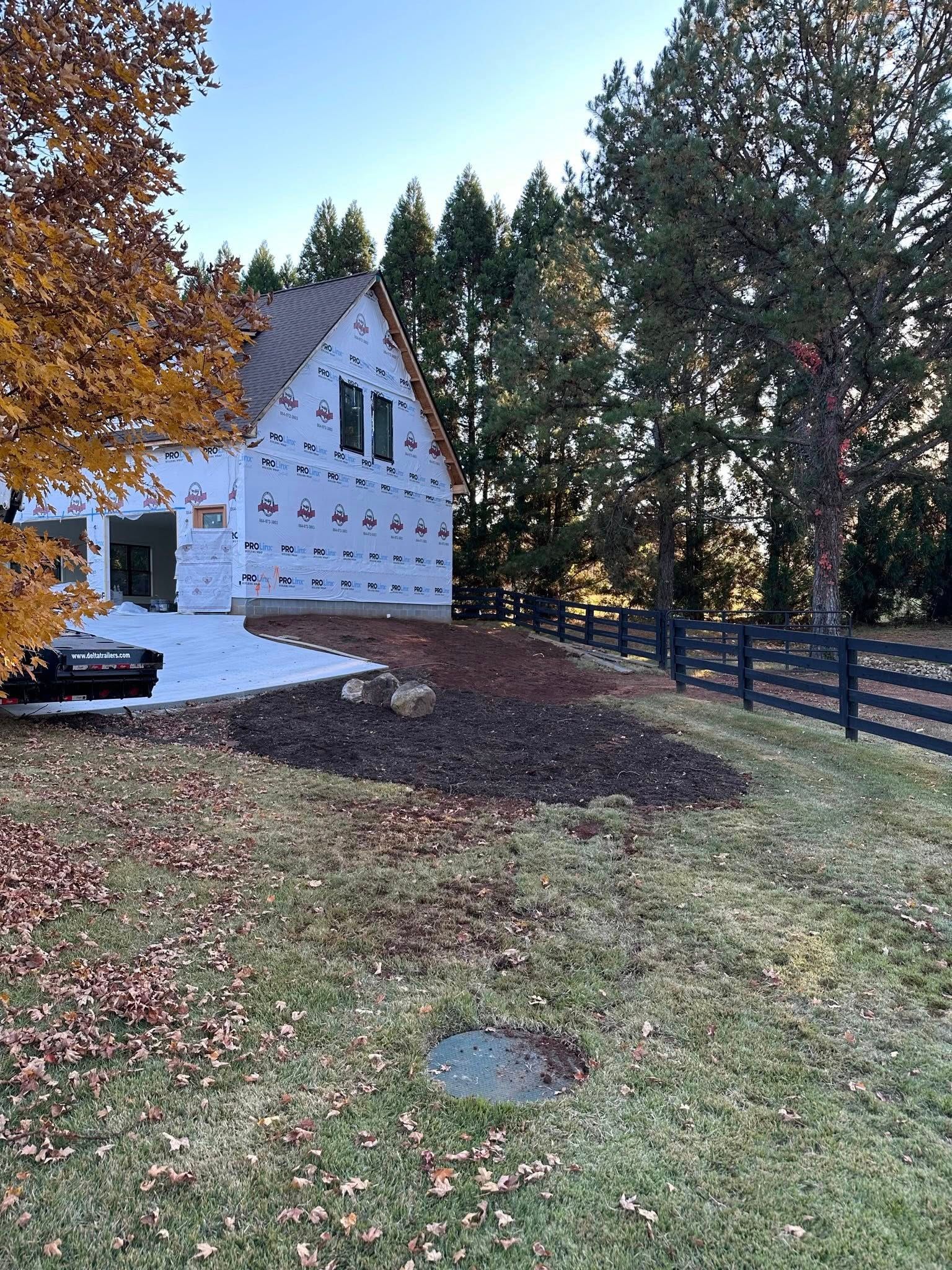 House under construction, blue wrap. Yard with brown mulch, black fence, and trees. Snow on the ground.