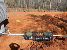 A black plastic septic tank is partially buried in red soil next to a house with a white pipe connecting to it.