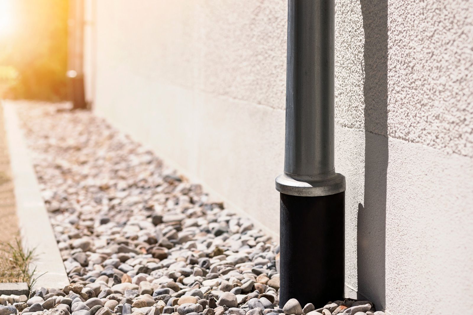 Black downspout along a light exterior wall beside a rock strip, lit by warm sunlight