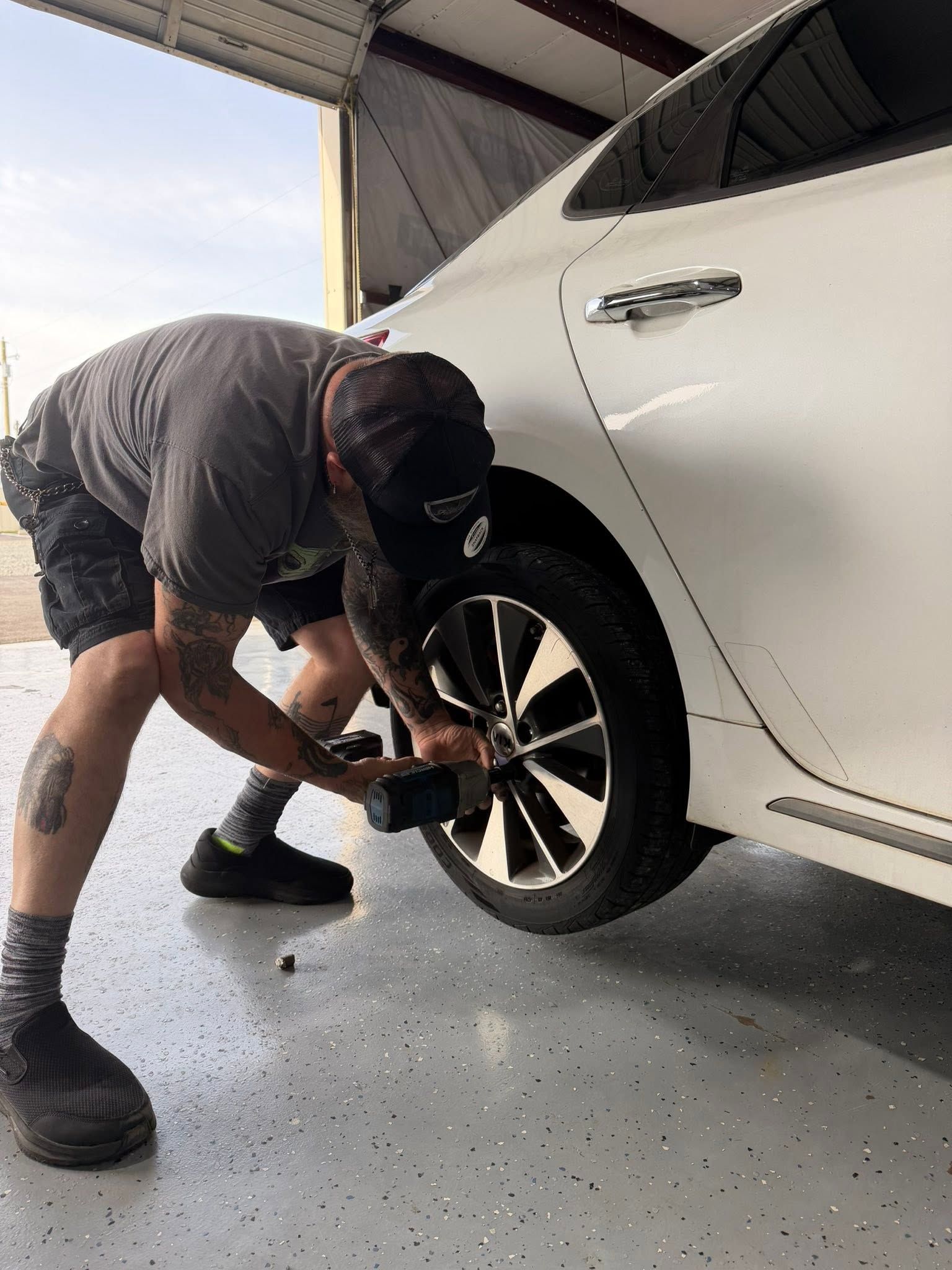 Person crouches using a power tool to remove a wheel from a white car in a garage.