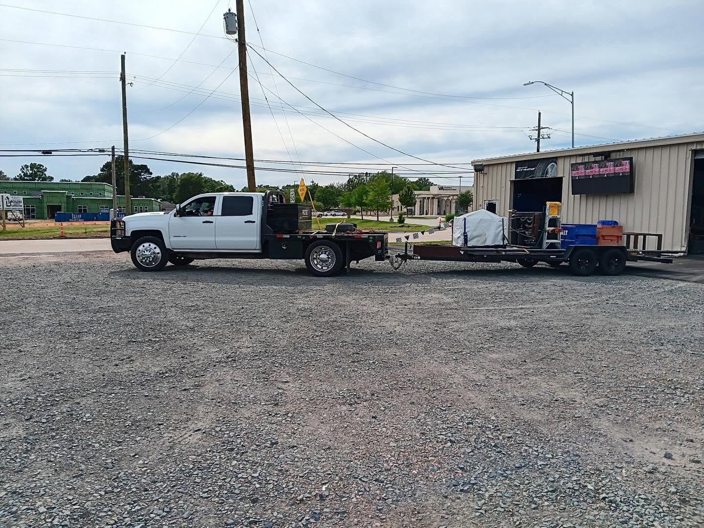 White pickup truck towing a flatbed trailer beside a small roadside building on gravel lot