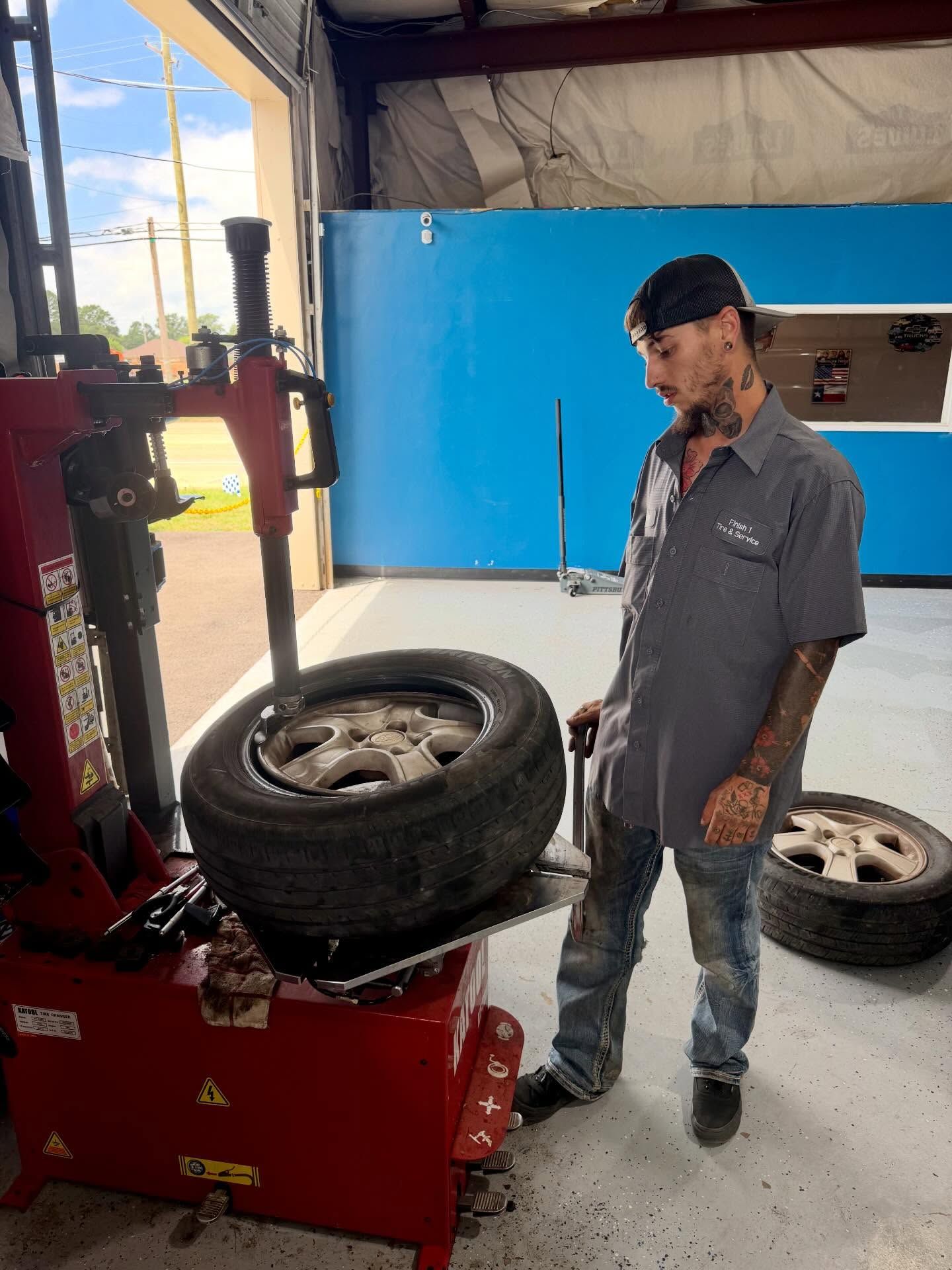 Mechanic standing beside a tire-changing machine in a garage, with tires on the machine and floor.