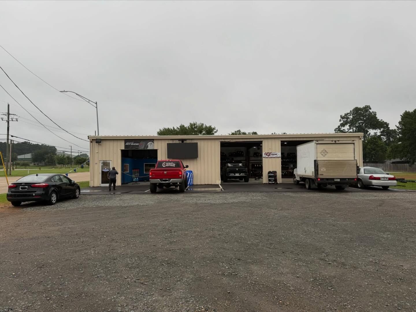 Gravel lot with a small auto shop, several parked vehicles, and an overcast sky