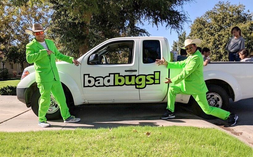 Two men in green suits are standing in front of a truck that says BadBugs!