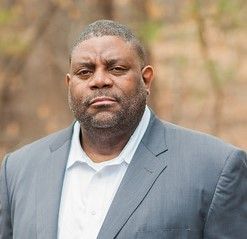A man in a suit and white shirt is standing in front of trees.