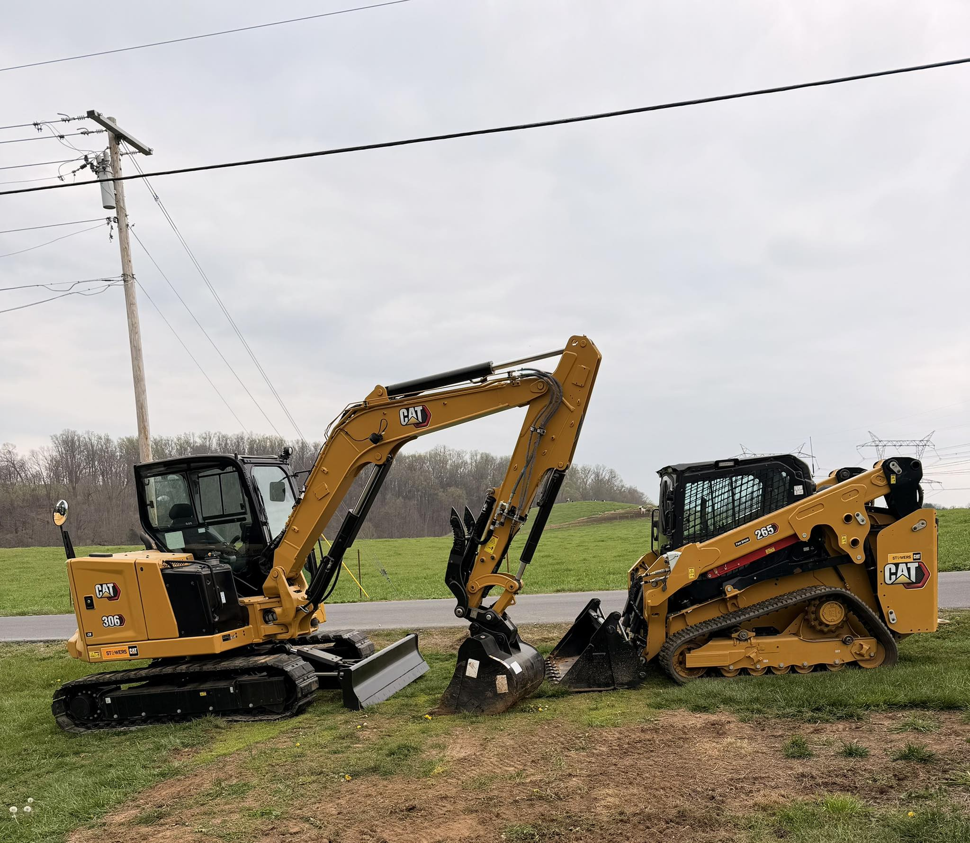 Two yellow excavators face each other in a grassy field beside power lines.