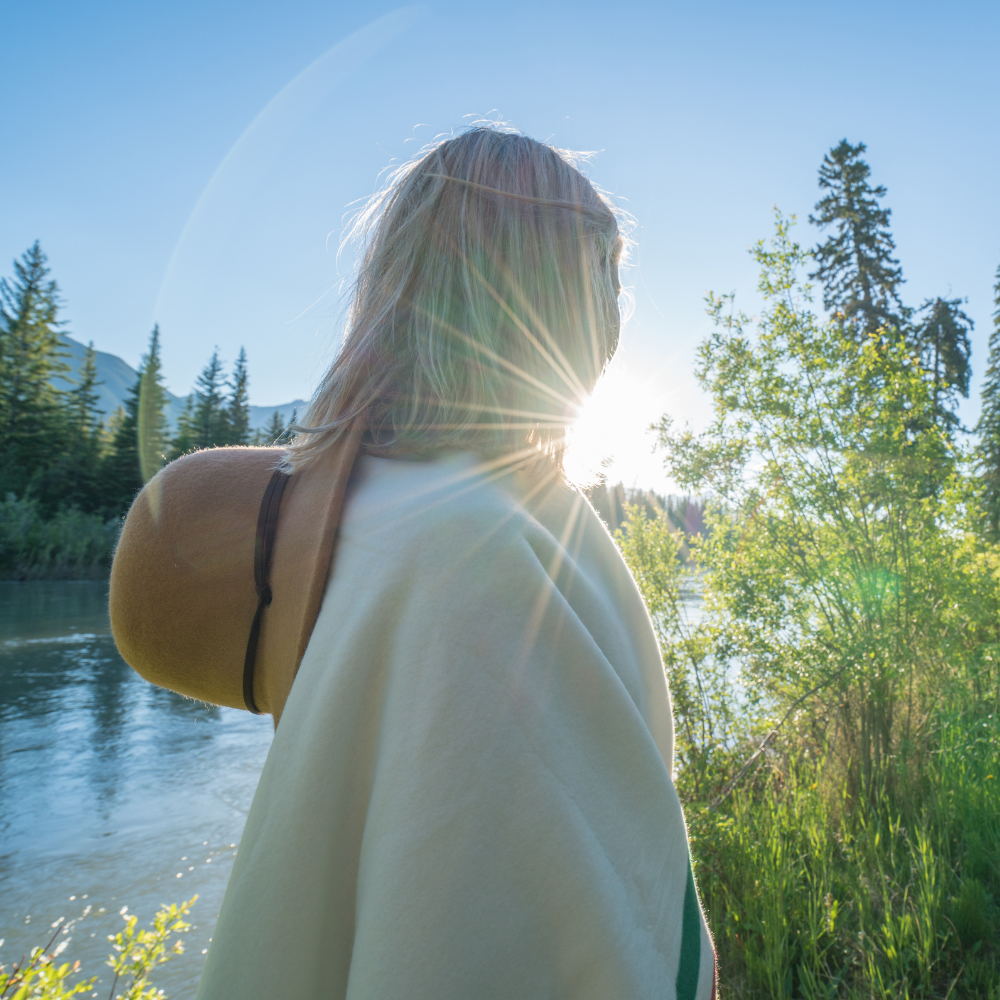 Person by a river, sun shining behind them, wearing a hat and light-colored shawl, trees in the background.