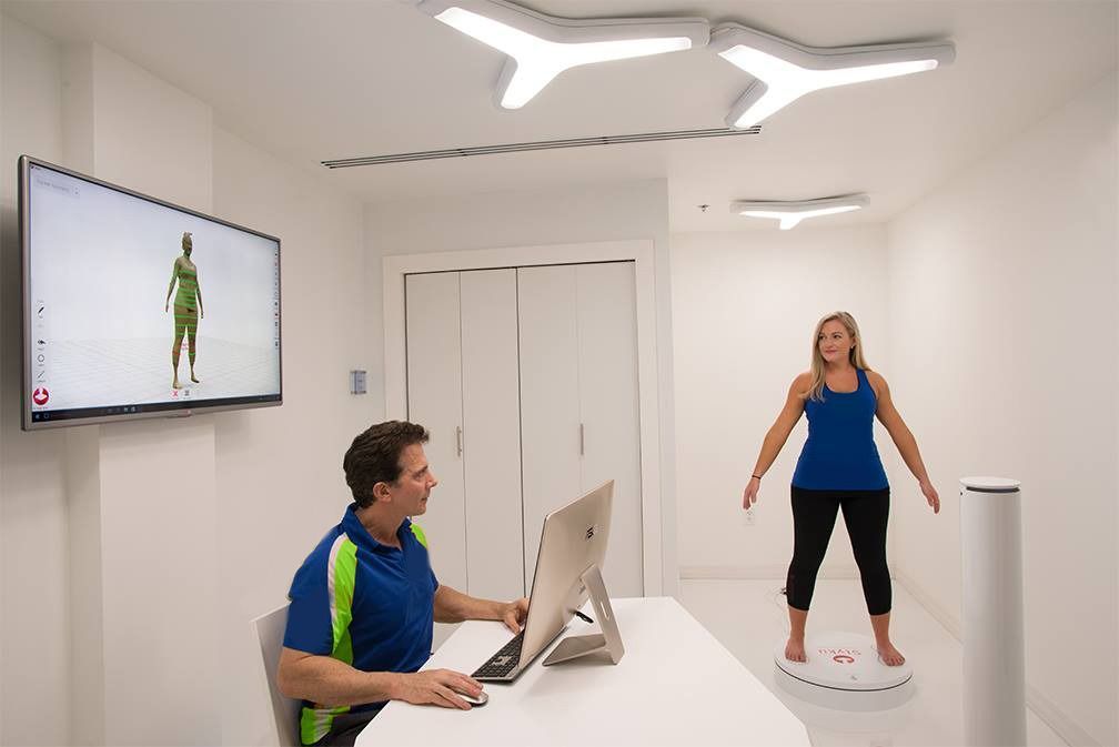 Man operating computer, woman standing on a scale, and a screen displays a figure. Interior, white walls.
