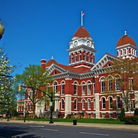 Red brick courthouse with arched windows, entrance under a portico, and large trees.