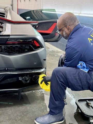 Mechanic inspecting the rear of a silver sports car in a garage, using a flashlight underneath.