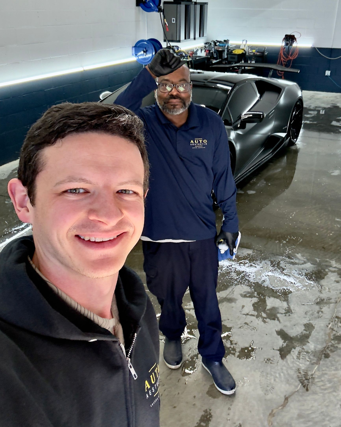 Two people smiling in a garage beside a black sports car, with tools and a wet floor in the background