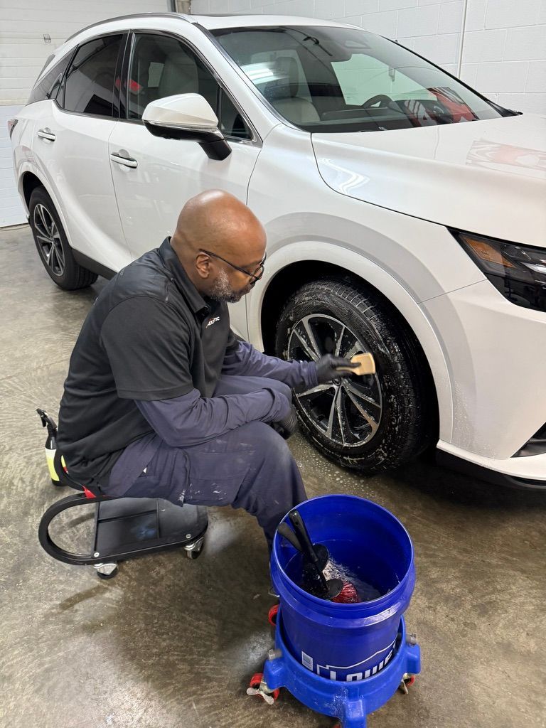 A professional detailer cleans the wheel of a white SUV while seated on a rolling stool next to a blue cleaning bucket.